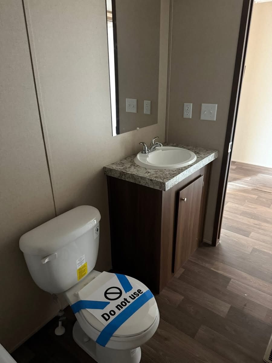 Bathroom with hardwood floor, single sink, and a mirror. Toilet has a blue "Do not use" tape. Neutral tones create a clean, restricted feel.