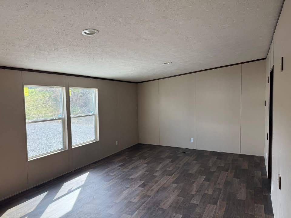 An empty room with beige walls and dark wood flooring. Two large windows let in natural light, creating a bright, open space. The tone is neutral and clean.