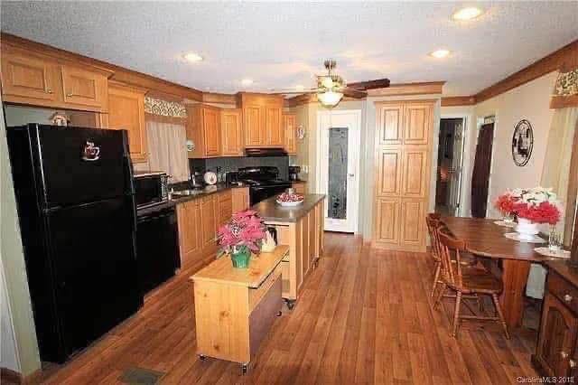 Warm kitchen with wooden cabinets, center island with flowers, and black appliances. Dining table with chairs on the right, ceiling fan above. Cozy ambiance.