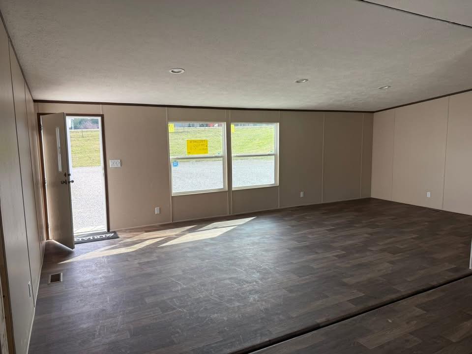 Spacious, empty room with wooden flooring, beige walls, and large windows letting in sunlight. The open door reveals a grassy landscape outside.