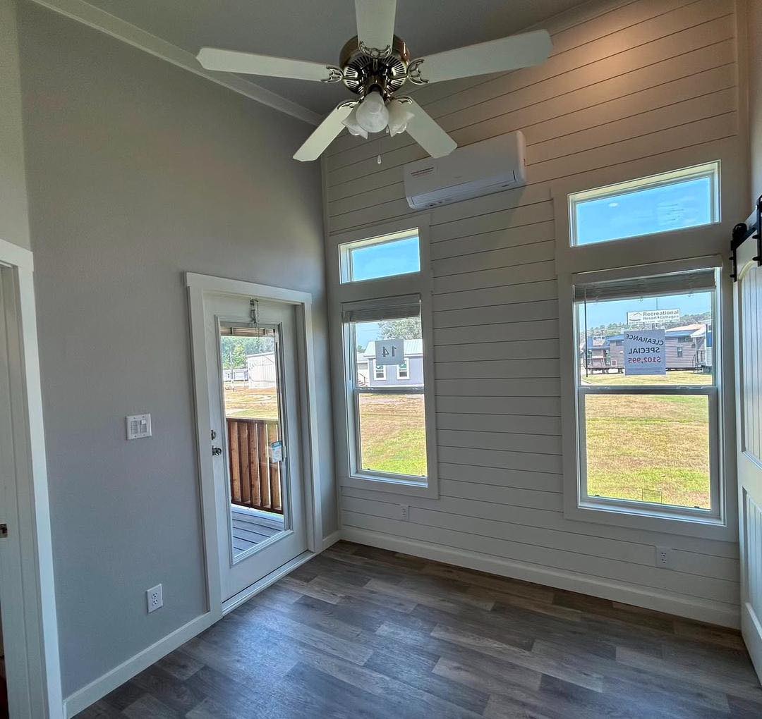 A small room with a high ceiling, featuring a white fan, light gray walls, and wood flooring. Three tall windows and a glass door offer outdoor views.