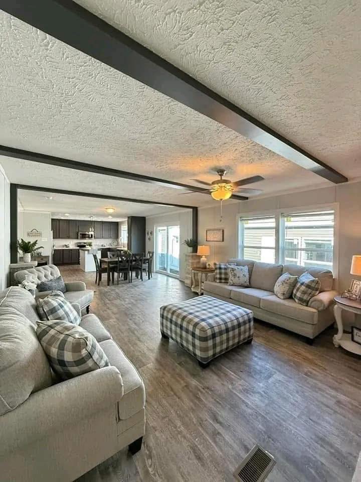 Spacious living room with neutral-toned sofas and plaid cushions, a matching ottoman, wood flooring, ceiling fan, and open kitchen dining area in the background.