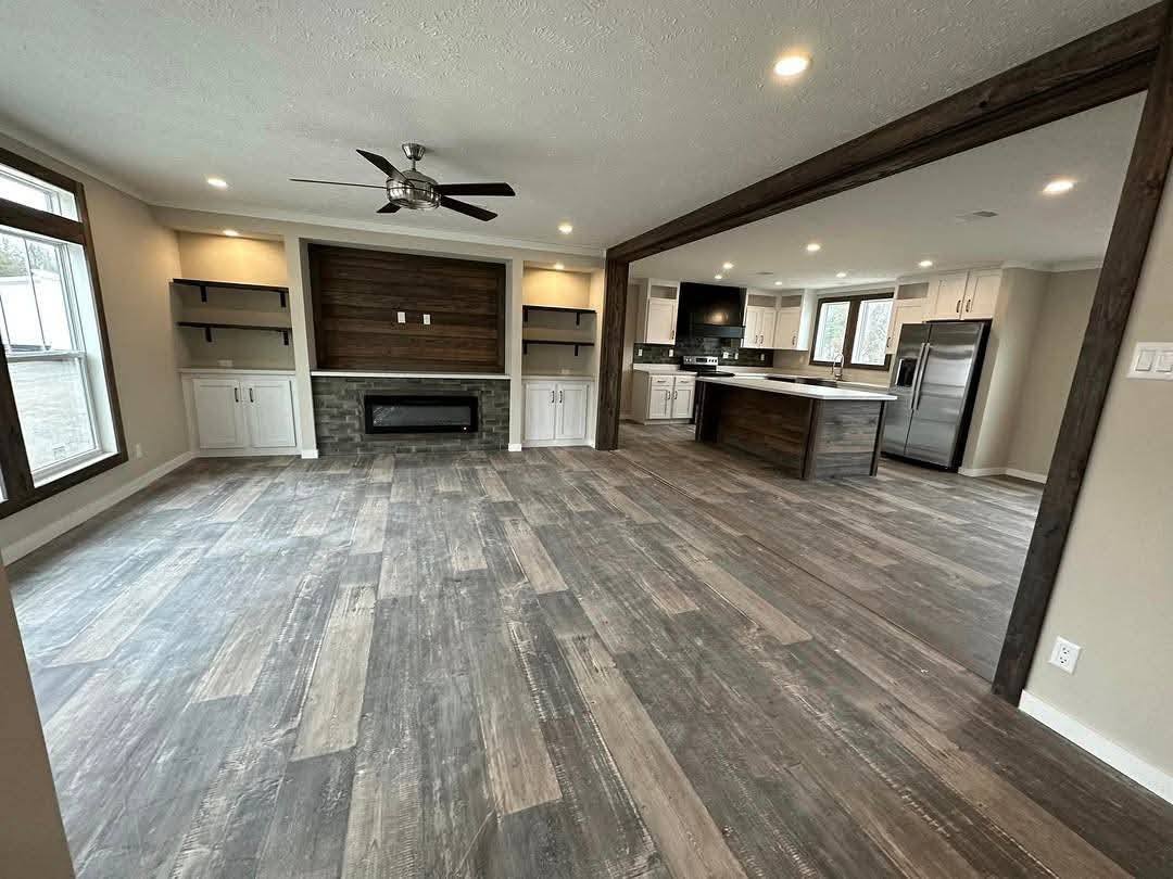 Spacious open-concept living room and kitchen with rustic wood floors, modern fireplace, ceiling fan, and dark wood trim, creating a cozy yet modern vibe.