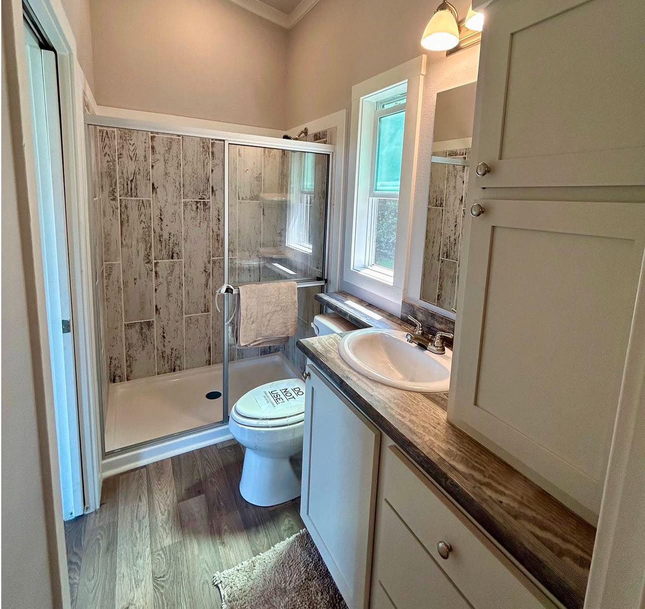 Small bathroom with a wood-patterned shower enclosure, glass sliding doors, and a white toilet. A sink with a marble countertop is beside a window. Cozy and clean atmosphere.