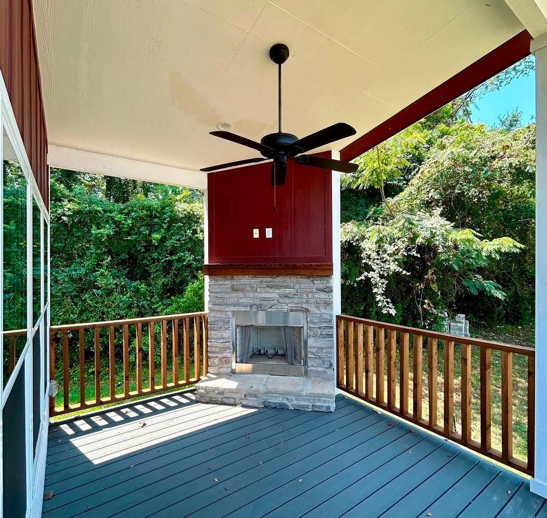 Covered porch with a ceiling fan and stone fireplace, surrounded by a wooden railing. Lush greenery in the background gives a serene, cozy feel.