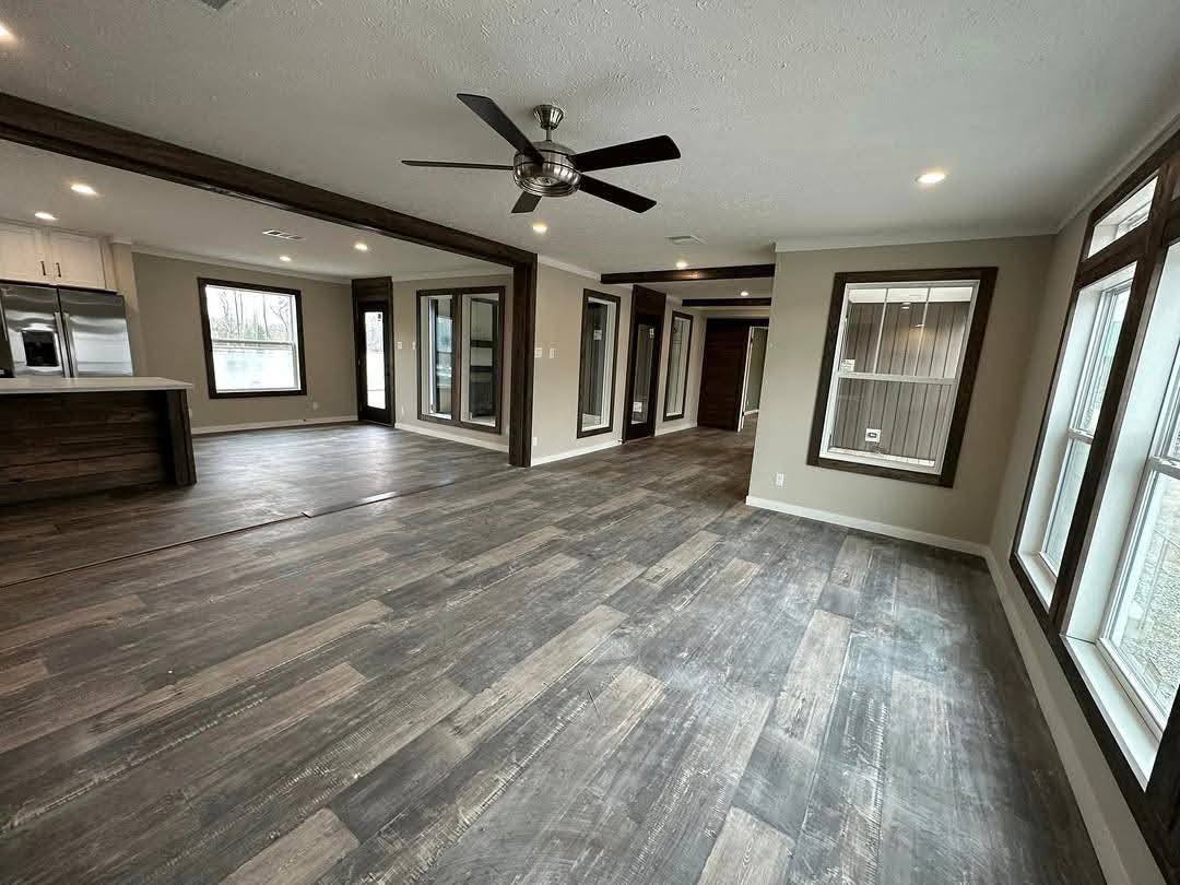 Spacious modern living area with dark wood flooring, ceiling fan, large windows, and recessed lighting. Open layout connects to a sleek kitchen.