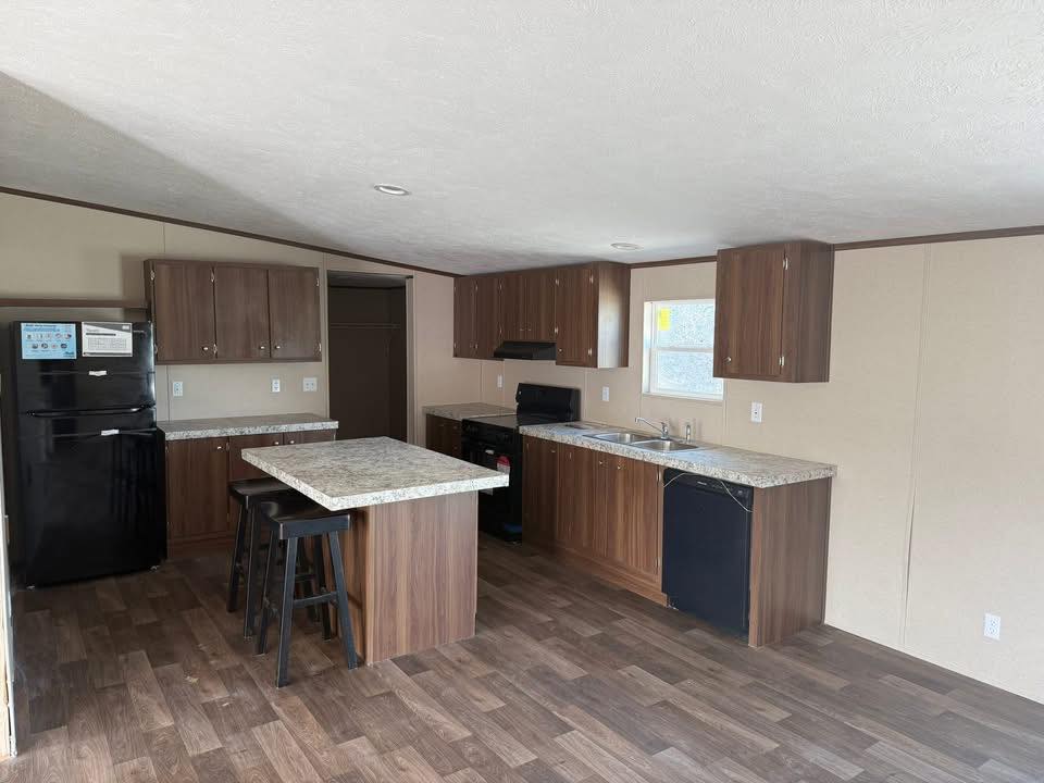 Modern kitchen with brown cabinets and marble countertops. Features a black fridge, oven, dishwasher, and a central island with stools on hardwood flooring.