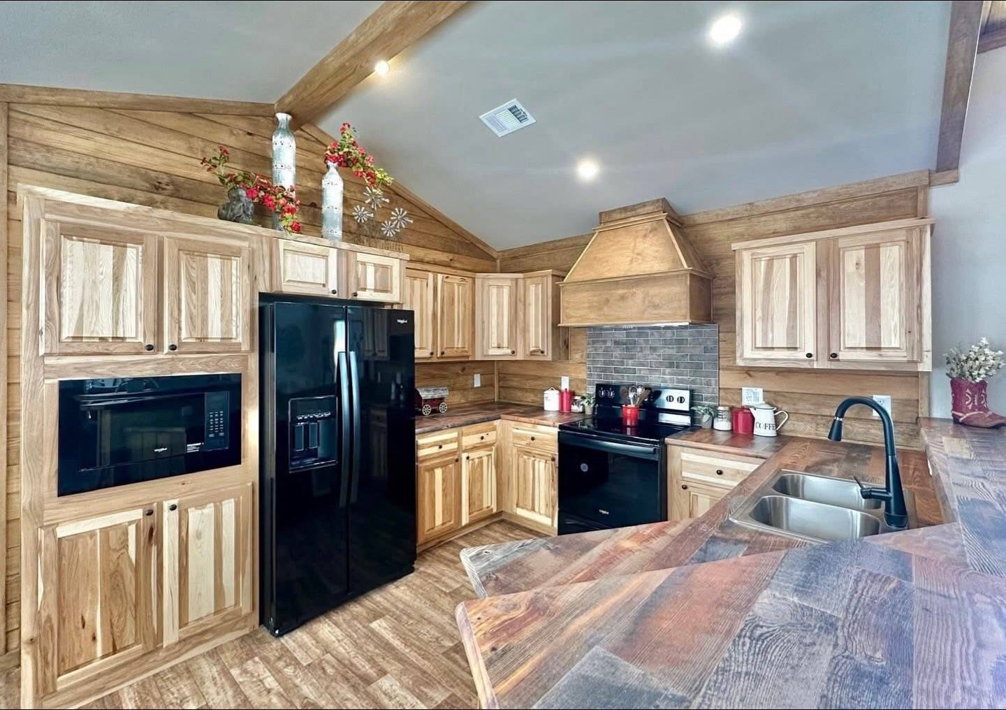 Rustic kitchen with wooden cabinets and a black fridge, stove, and microwave. Brick backsplash under a wooden hood. Red accents and floral decor. Warm, cozy ambiance.