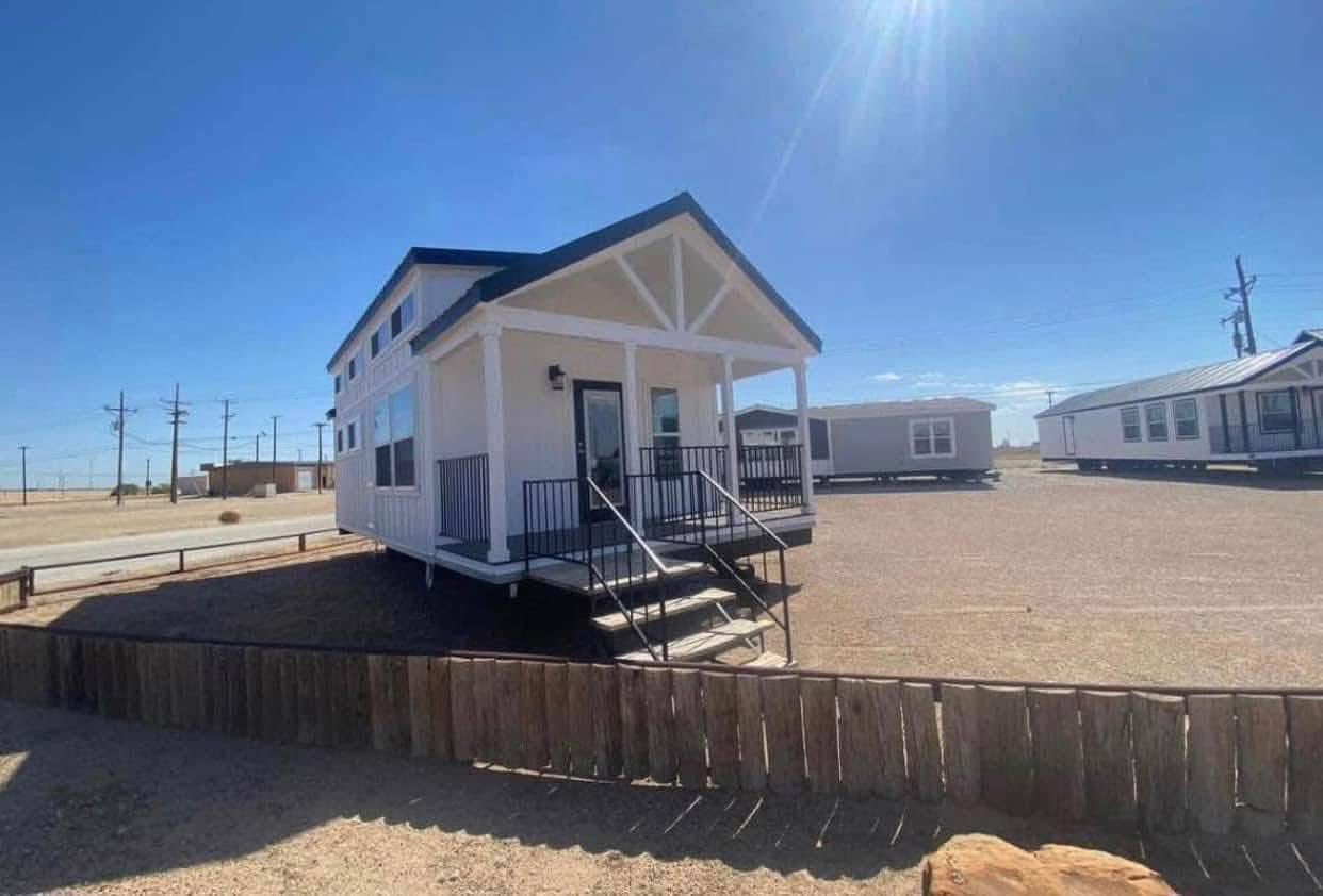 A small white house with a porch and black railings sits in a sunny, arid landscape with clear blue skies. Nearby, similar houses are visible.