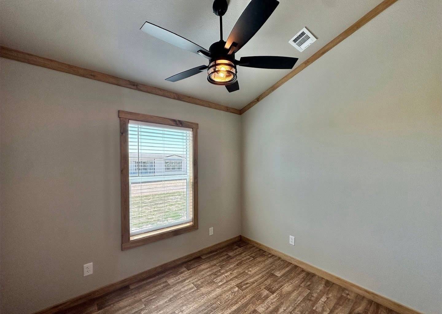 Small empty room with light gray walls, wood trim, and vinyl wood flooring. A ceiling fan with five black blades hangs centrally, and a single window allows natural light in.