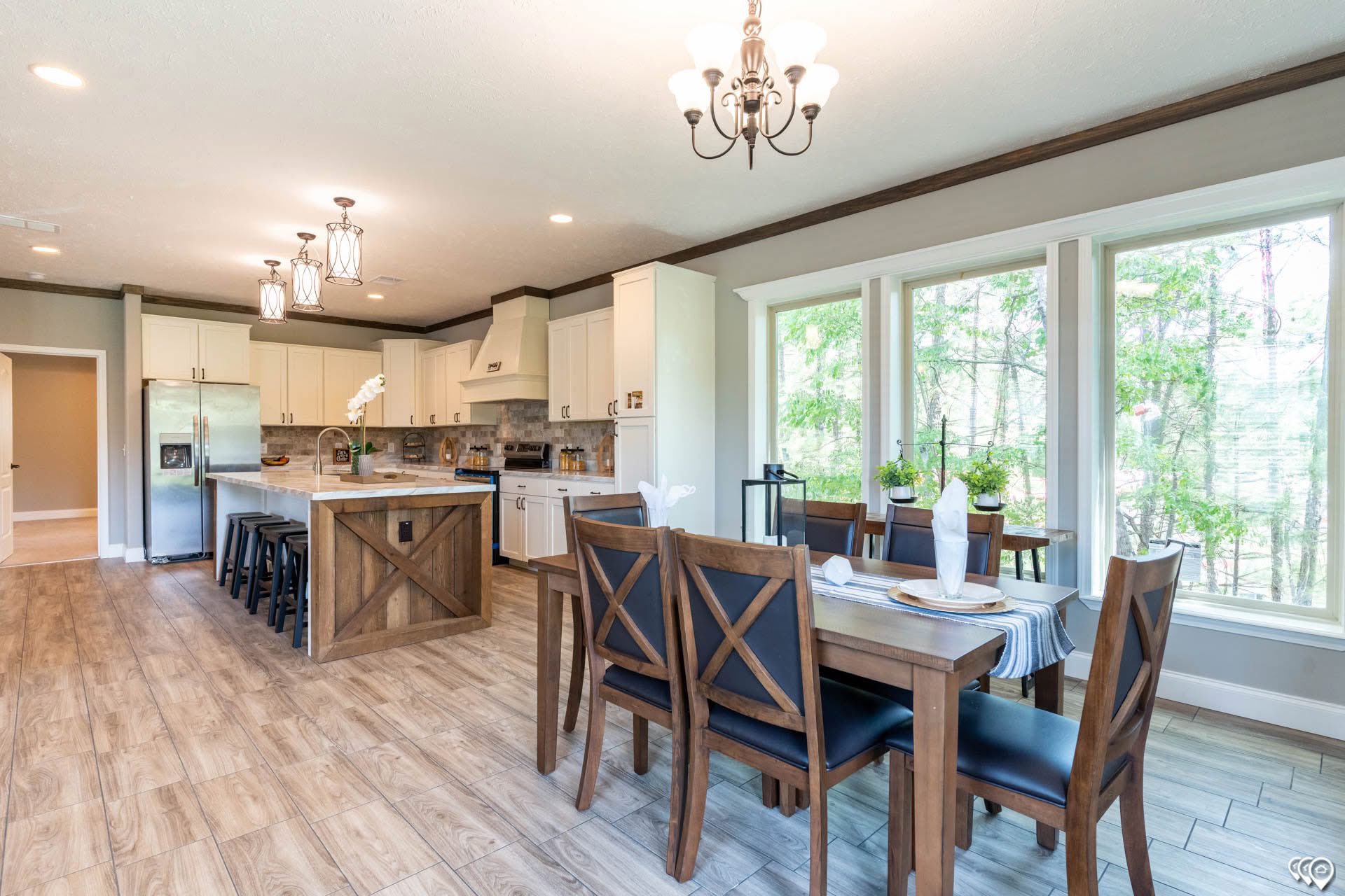 Spacious kitchen-dining area with wooden floors. Modern kitchen features an island with bar stools, stainless steel appliances, and pendant lights. Adjacent dining table set by large windows overlooking greenery. Cozy and inviting atmosphere.