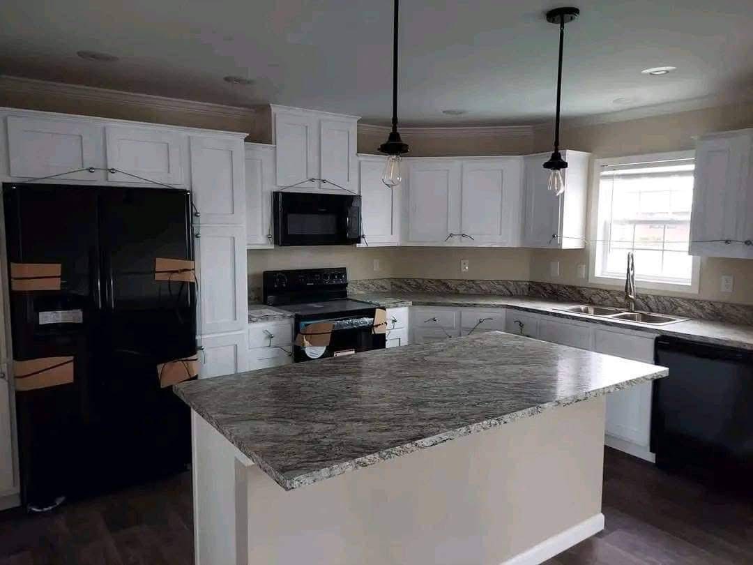 Modern kitchen with white cabinets, granite countertops, dark appliances, and pendant lights over the island. Bright with natural light from a window.