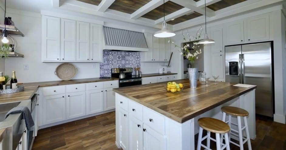 Spacious kitchen with white cabinets, wooden countertops, and a large island with stools. Features stainless steel fridge and patterned tile backsplash.
