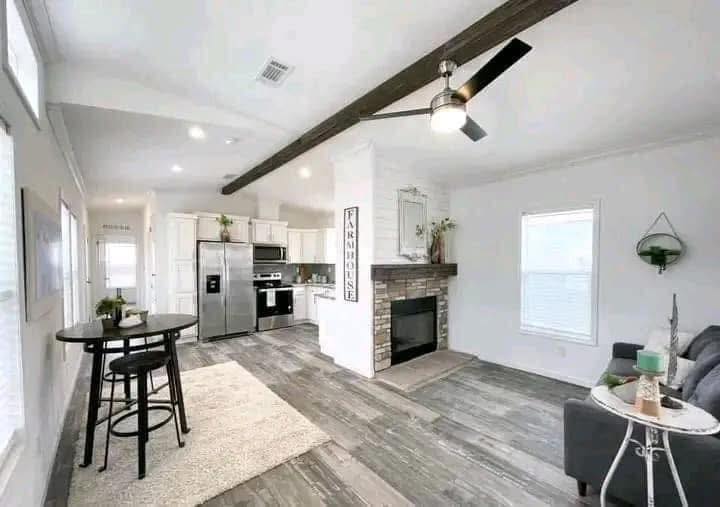 Bright open-concept living room and kitchen with rustic accents. Features a stone fireplace, ceiling fan, wood beam, and modern furnishings. Cozy and inviting.