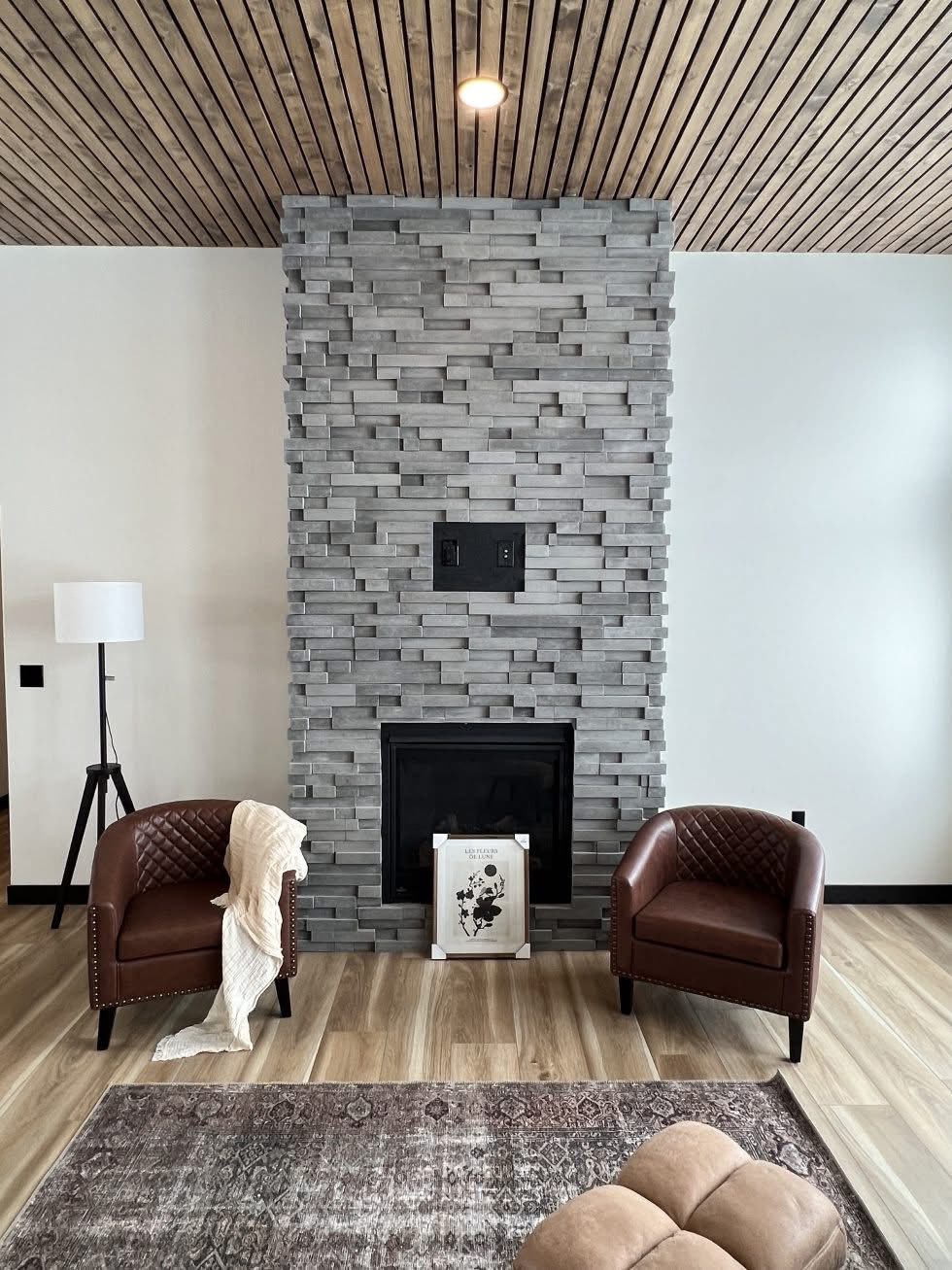 Modern living room with a stone fireplace, two brown leather chairs, and a cozy throw. The wooden ceiling and floor create a warm, inviting atmosphere.