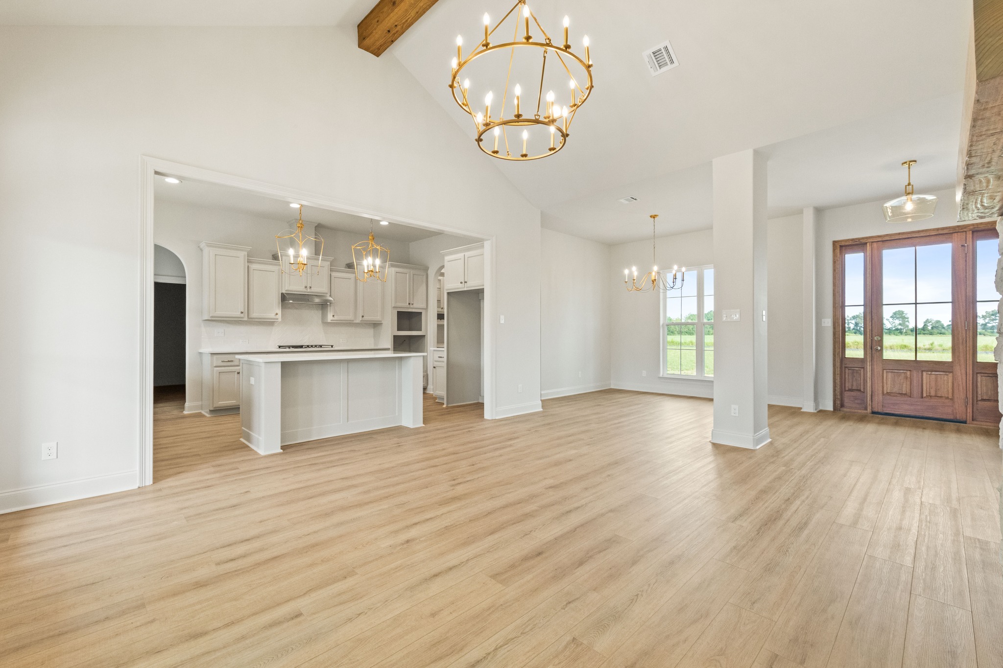Spacious open-plan kitchen and living area with light wooden floors, high ceilings, and elegant chandeliers. Large windows and wooden door offer scenic views.