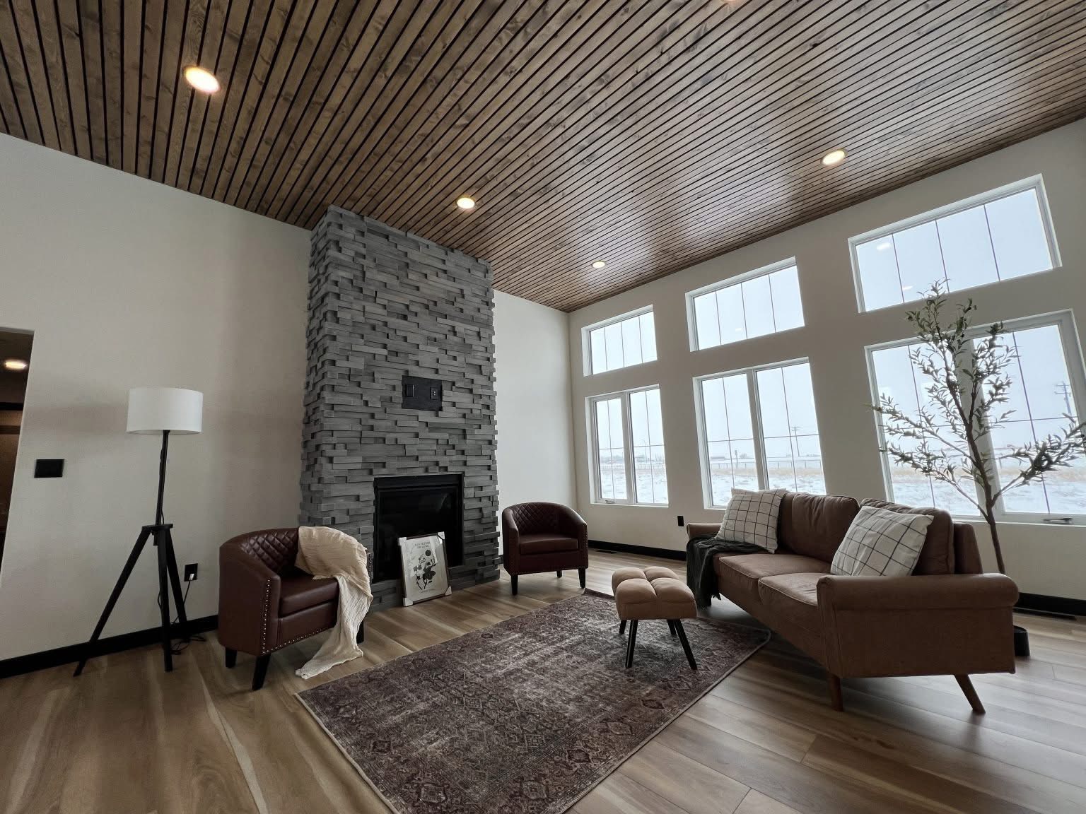 Modern living room with a stone fireplace, brown leather seating, and wooden ceiling. Large windows offer natural light, creating a cozy ambiance.