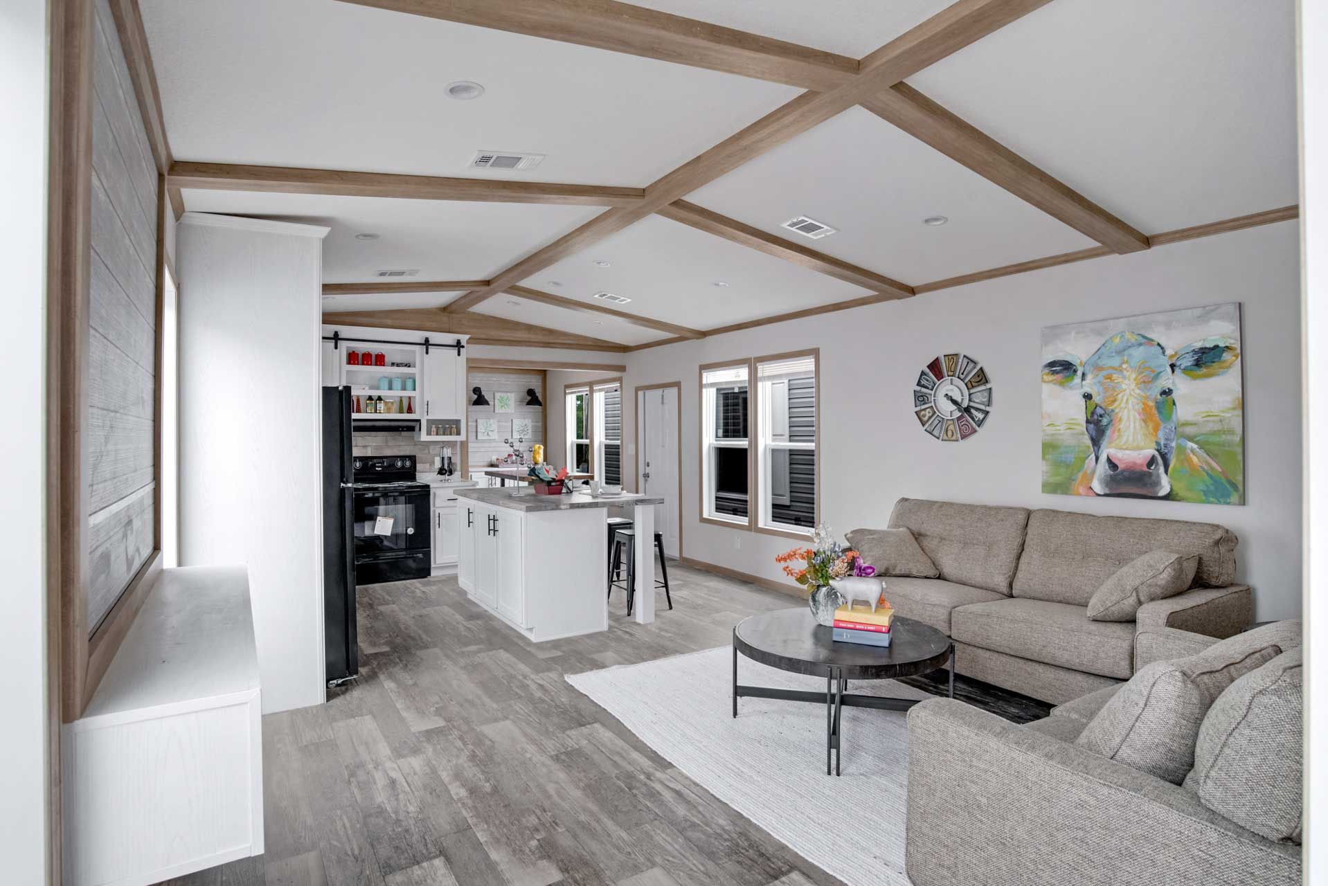 Modern open-concept living room and kitchen with wood beam ceiling. Gray sofas, cow painting, round coffee table with flowers, and kitchen island. Cozy and inviting.