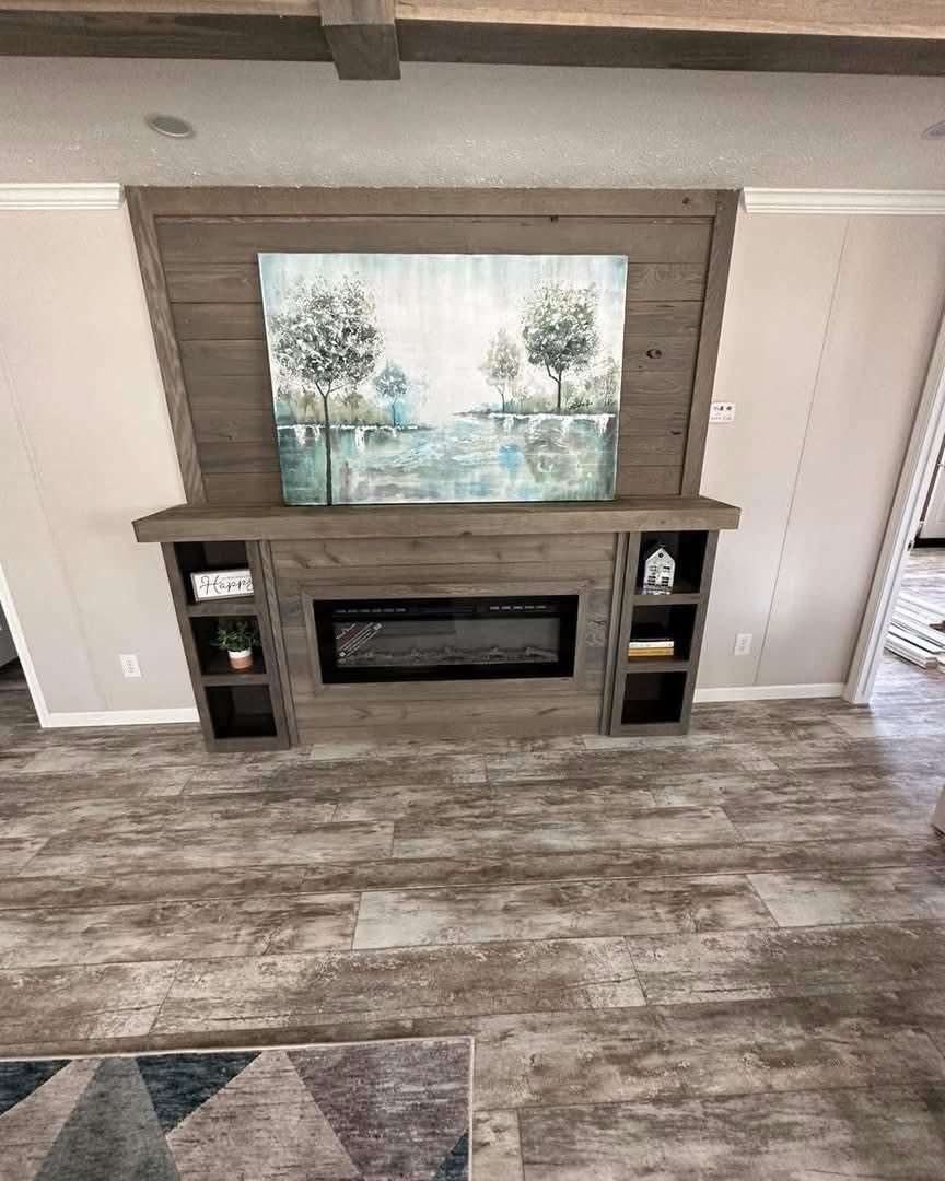 A cozy living room features a wooden fireplace with shelves, displaying small decorations. Above, a soothing landscape painting evokes a serene atmosphere.