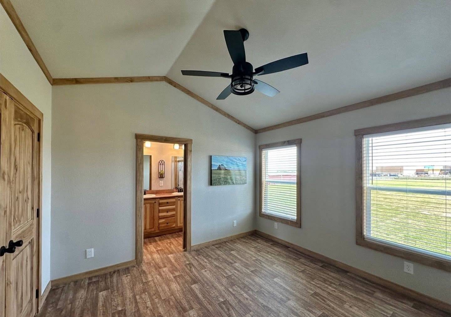 A spacious room with wood accents features two large windows with blinds, a ceiling fan, and an open doorway leading to a bathroom. Natural light creates a warm, inviting atmosphere.