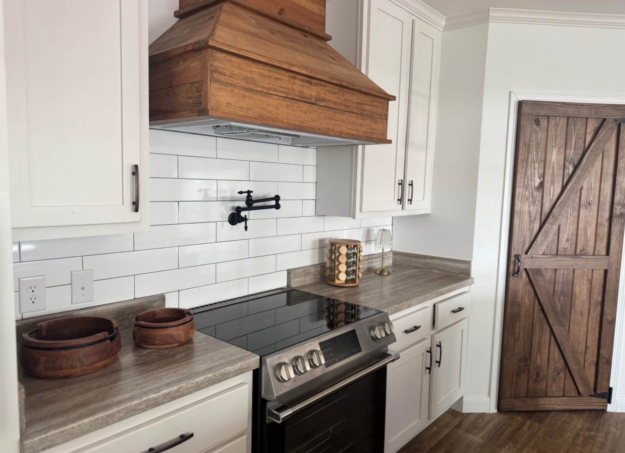 A rustic kitchen featuring white cabinets, a wooden range hood, subway tile backsplash, and a barn door. Cozy ambiance with a modern stove and wooden bowls.