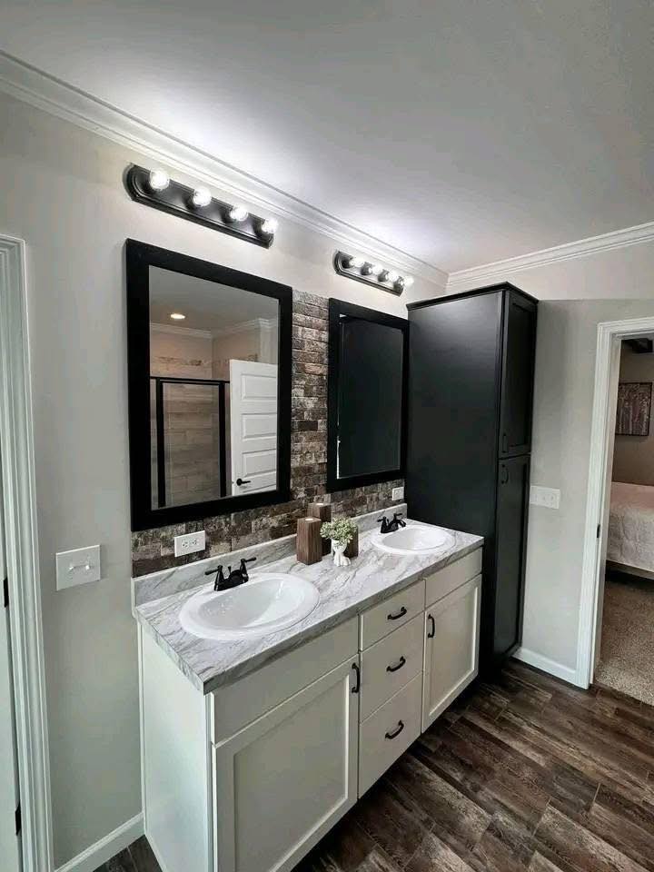 Modern bathroom with dual sinks on a granite countertop, each under a framed mirror. Warm lighting, dark cabinet, and wood-paneled floor create a cozy vibe.