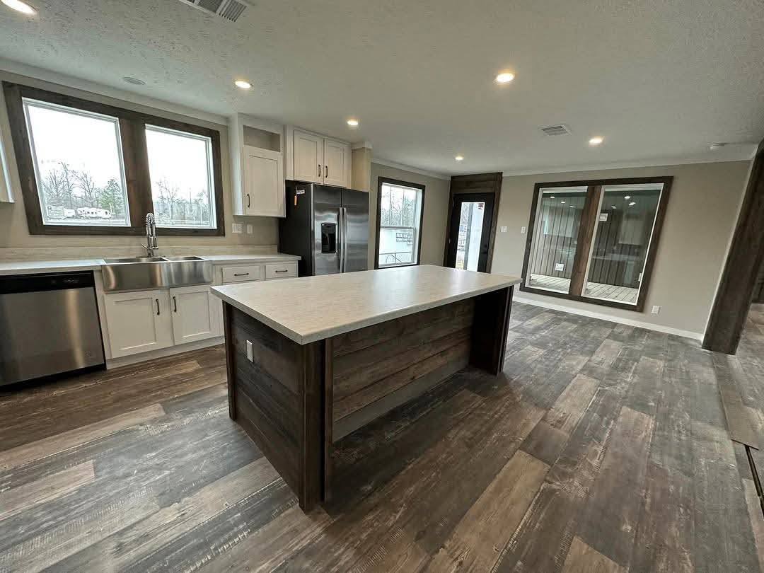 Modern kitchen with dark wood floors, a large central island with light countertop, white cabinets, stainless steel appliances, and large windows.