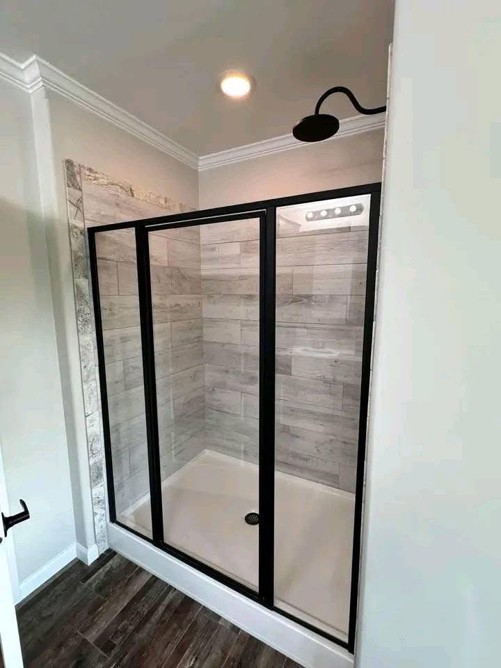 Modern shower with a glass door and black frame, featuring gray wood-look tiles. The design is sleek, with a dark shower head, exuding a minimalistic tone.