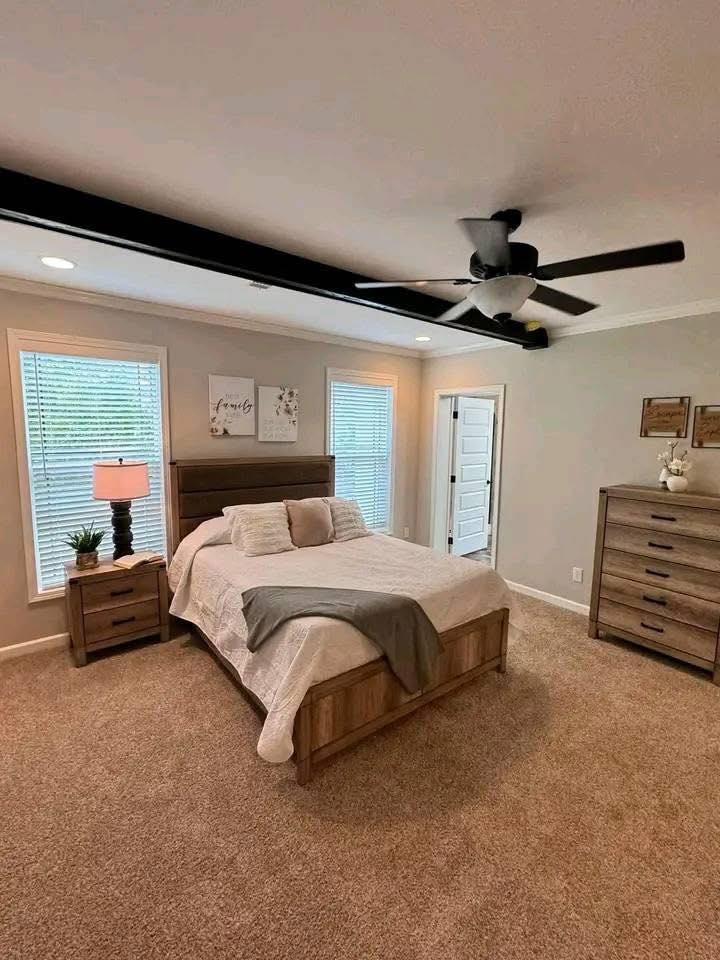 A cozy bedroom with a large bed centered, flanked by nightstands and lamps. A ceiling fan hangs above, and two windows let in natural light. Earth tones create a warm, inviting ambiance.