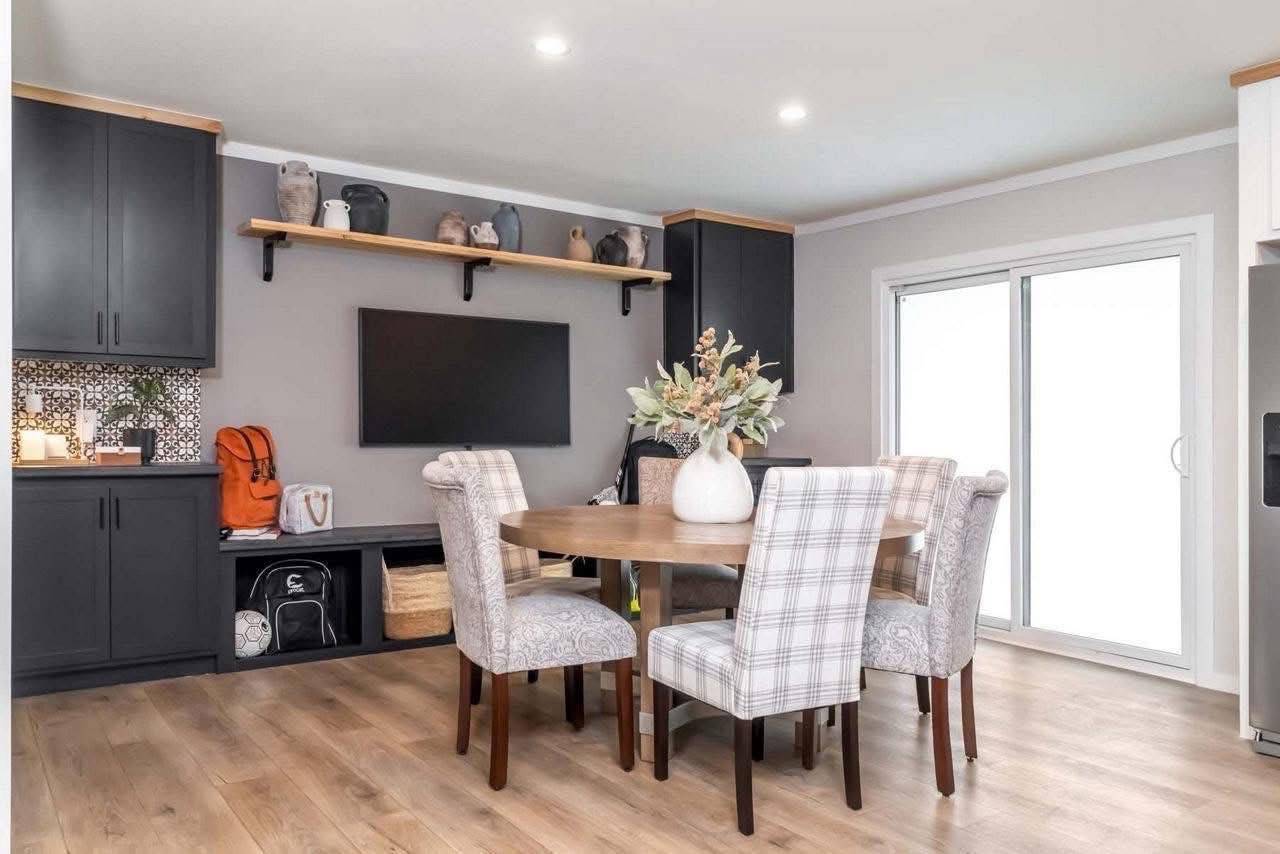 A modern dining area with a round wooden table and plaid upholstered chairs. Behind is a wall-mounted TV, black cabinets, and a shelf with pottery. A vase with flowers is on the table, and large sliding doors allow natural light in, creating a warm, inviting atmosphere.