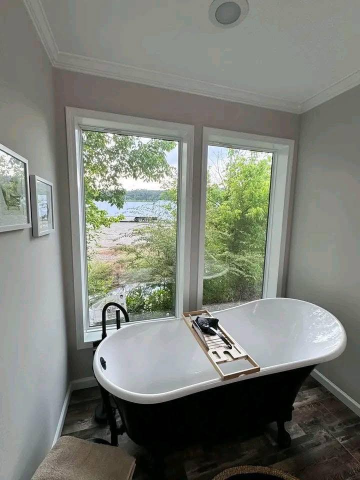 Freestanding bathtub by large windows with a view of lush greenery and a serene lake. Soft lighting emphasizes a peaceful, relaxing atmosphere.