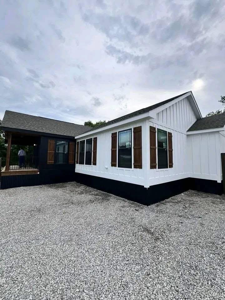 Single-story white house with dark trim, wooden shutters, and a gravel driveway. Overcast sky above adds a calm, subdued atmosphere.