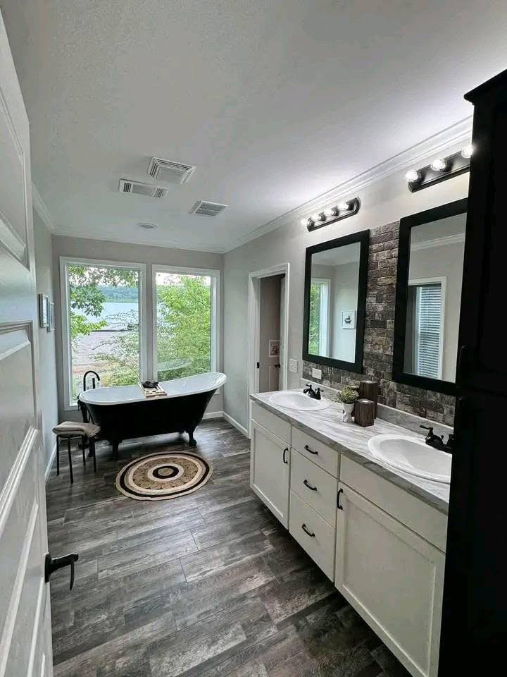 Modern bathroom with wood flooring, featuring a black freestanding tub by large windows and a double vanity with two framed mirrors. Bright, tranquil ambiance.