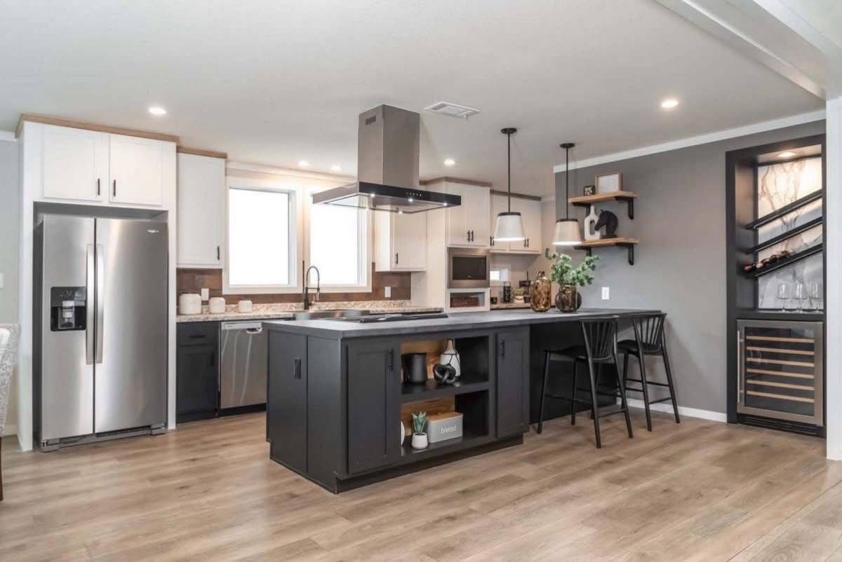 Modern kitchen with wooden floors, featuring a central island with black cabinets, pendant lights, and bar stools. Stainless steel appliances and a wine rack add a sleek touch.