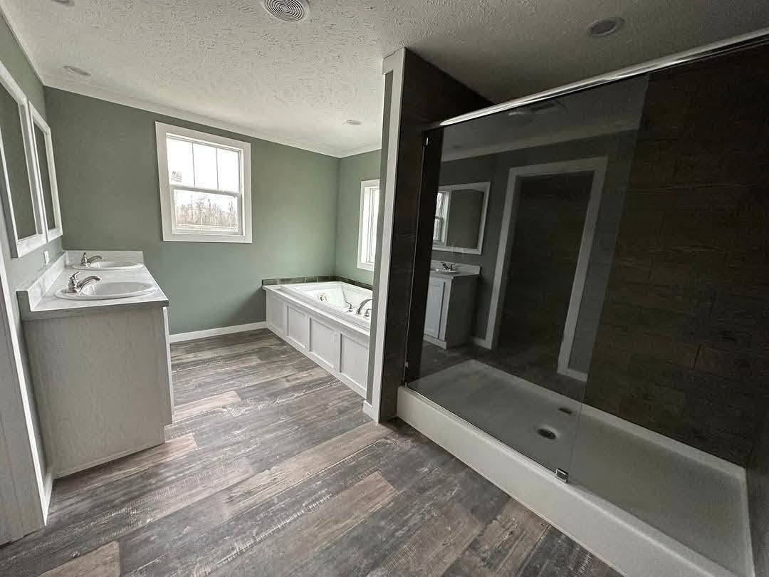 Spacious bathroom with green walls, wood flooring, a double vanity, large mirror, and bathtub under a window. A glass-enclosed shower adds modernity.
