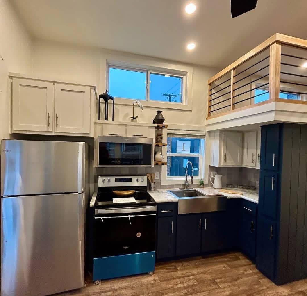 Modern kitchen with stainless steel appliances, navy and white cabinets, and a loft. Warm lighting, wooden accents, and a large farmhouse sink. Cozy ambiance.