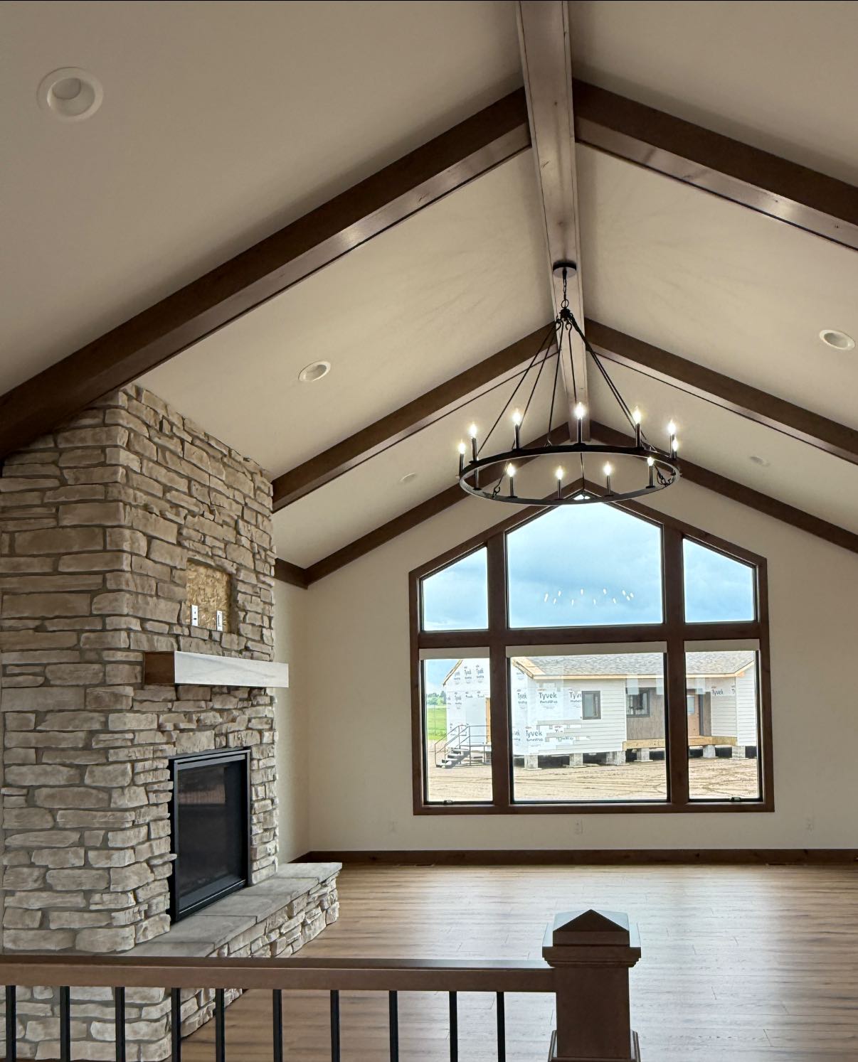 Spacious living room with vaulted ceiling, large stone fireplace, and expansive windows. A modern chandelier hangs, creating a cozy, open atmosphere.