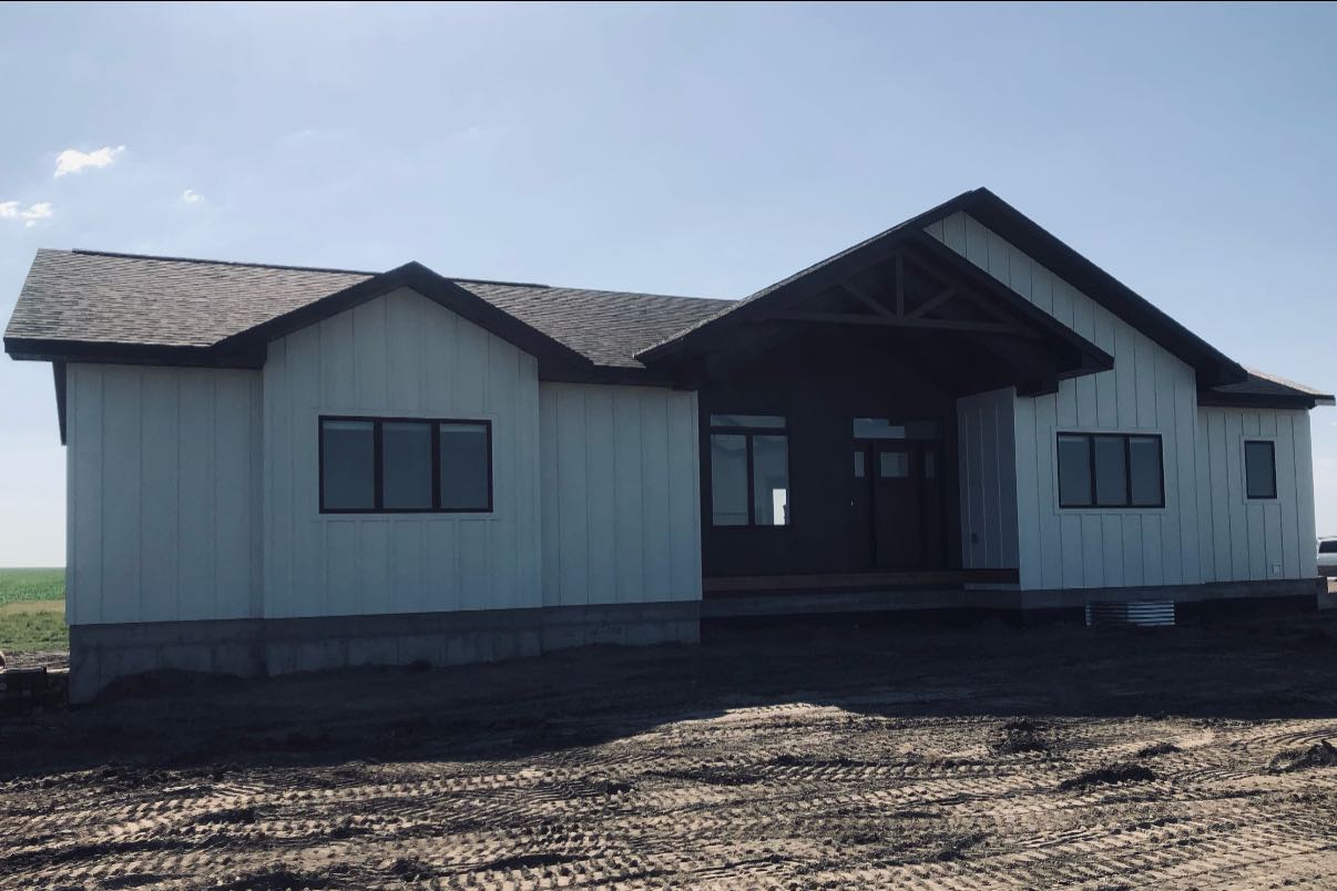 A single-story house with white paneling and a dark roof stands on bare earth under a clear sky. It has large windows and a centered front entrance.