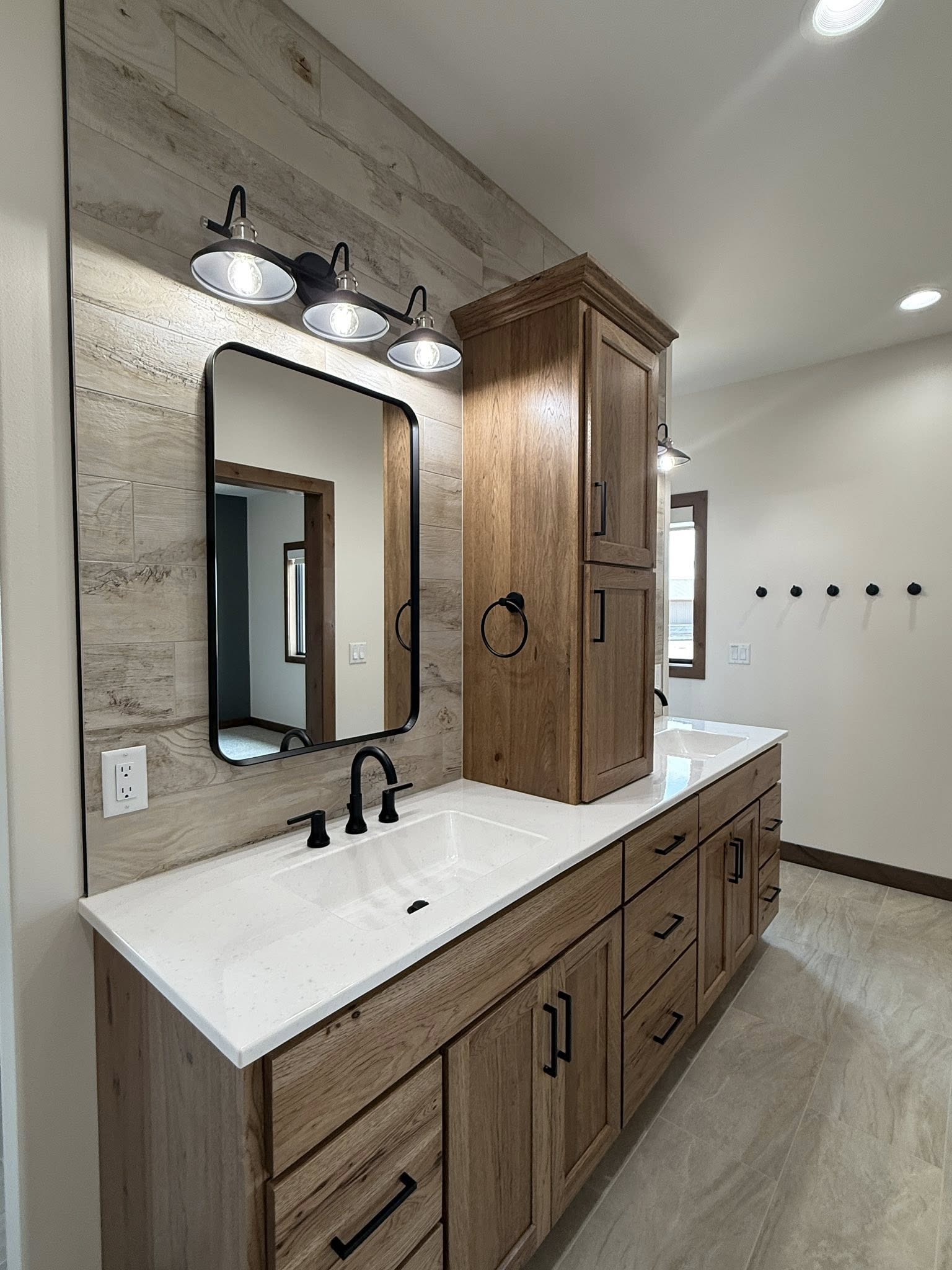 A modern bathroom vanity with a light wood finish, featuring a white countertop with two sinks. Above, a rectangular mirror is flanked by black fixtures.