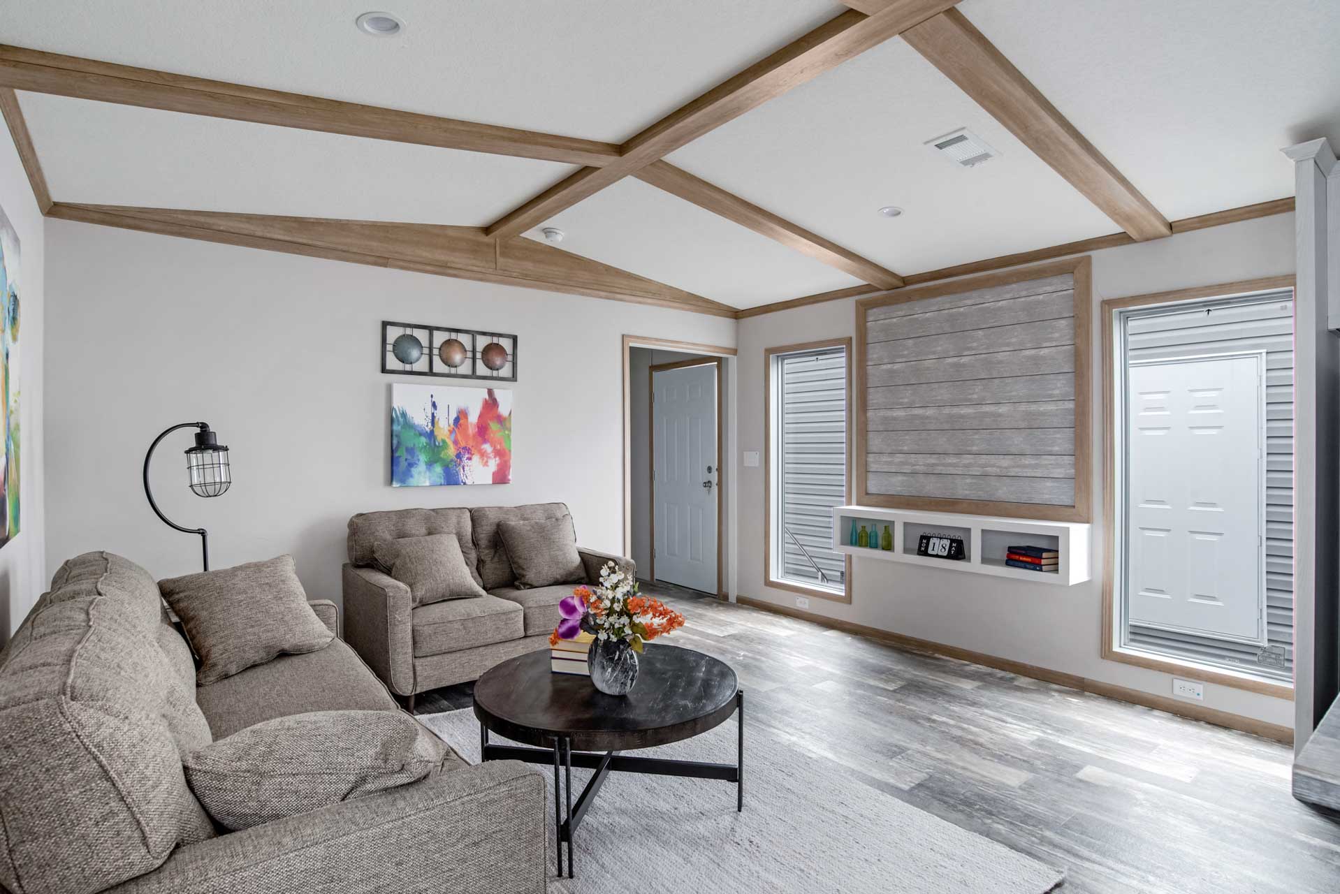 Modern living room with gray sofas, colorful artwork, and a black round coffee table with flowers. Light streams through large windows, creating a bright, inviting atmosphere.
