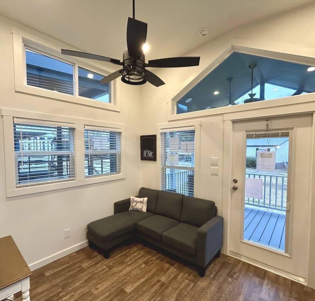 Bright living room with high ceiling features a dark gray L-shaped sofa, decorative cushions, wood flooring, and large windows. A ceiling fan adds a modern touch.