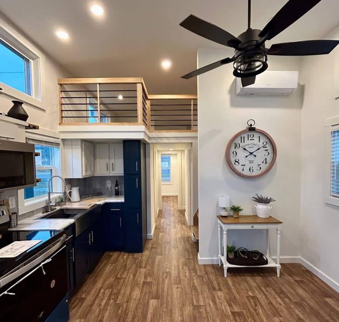 Modern kitchen and hallway with wooden floors, navy cabinets, and a loft. A large wall clock and ceiling fan add charm and a cozy, inviting feel.