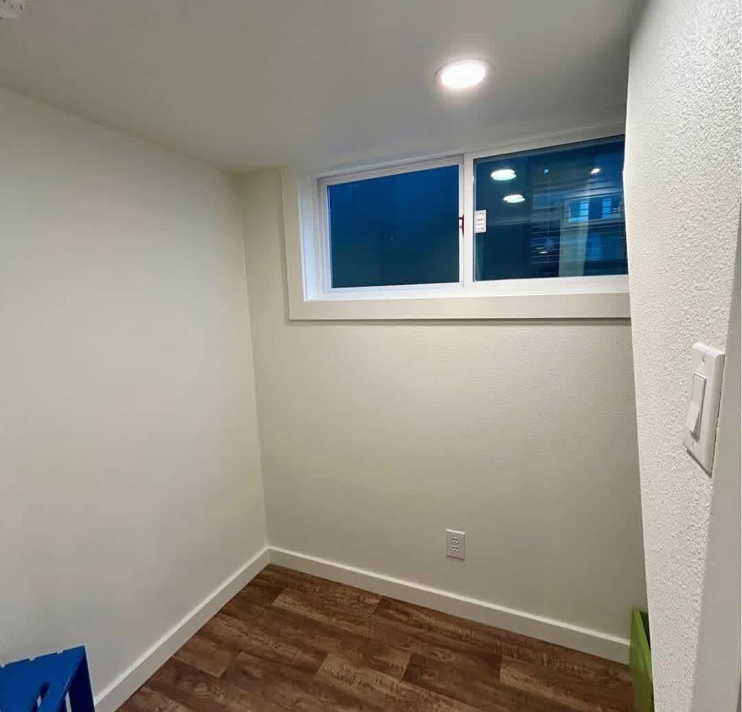 Small, empty room with white walls, a wood floor, and a narrow window near the ceiling. A single recessed light brightens the minimalist space.