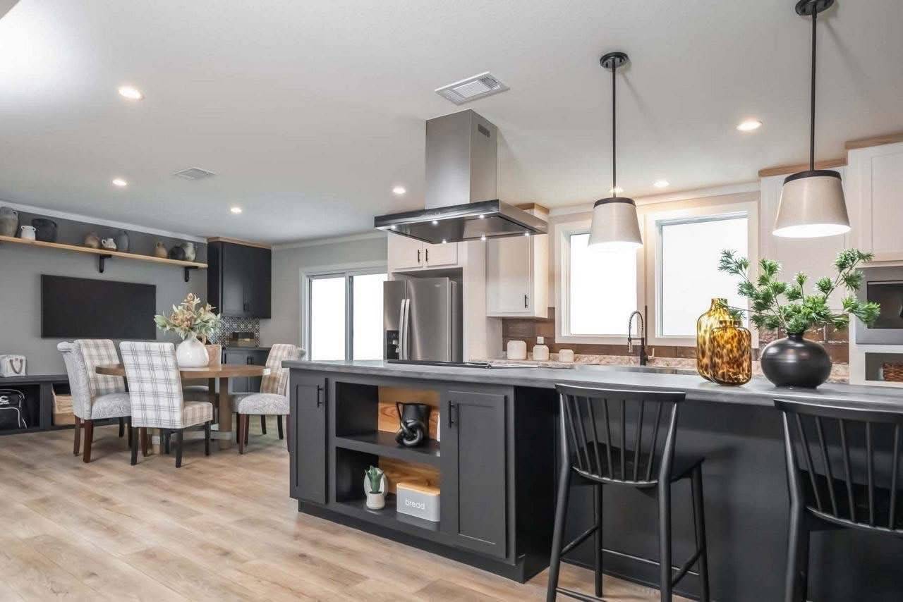 Modern kitchen with a black island, barstools, pendant lights, and stainless steel appliances. A cozy dining area with checkered chairs is in the background.