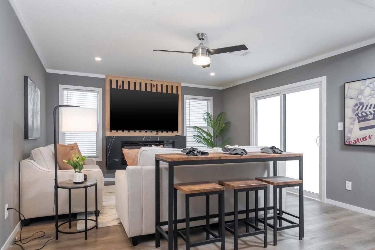 Modern living room with gray walls, a ceiling fan, and a large TV mounted on a wooden panel. Includes a light sofa, table with stools, and sliding doors.