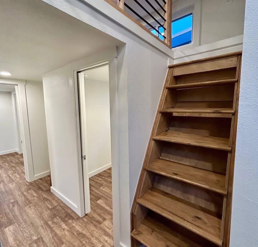 A narrow wooden staircase leads to a loft. The space below features white walls and wood-patterned flooring, with an open door to a bright room.