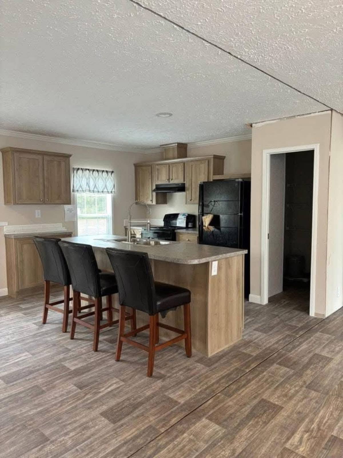 A modern kitchen features light wood cabinets, a black fridge, and a central island with three black padded chairs, set on grey tile flooring.