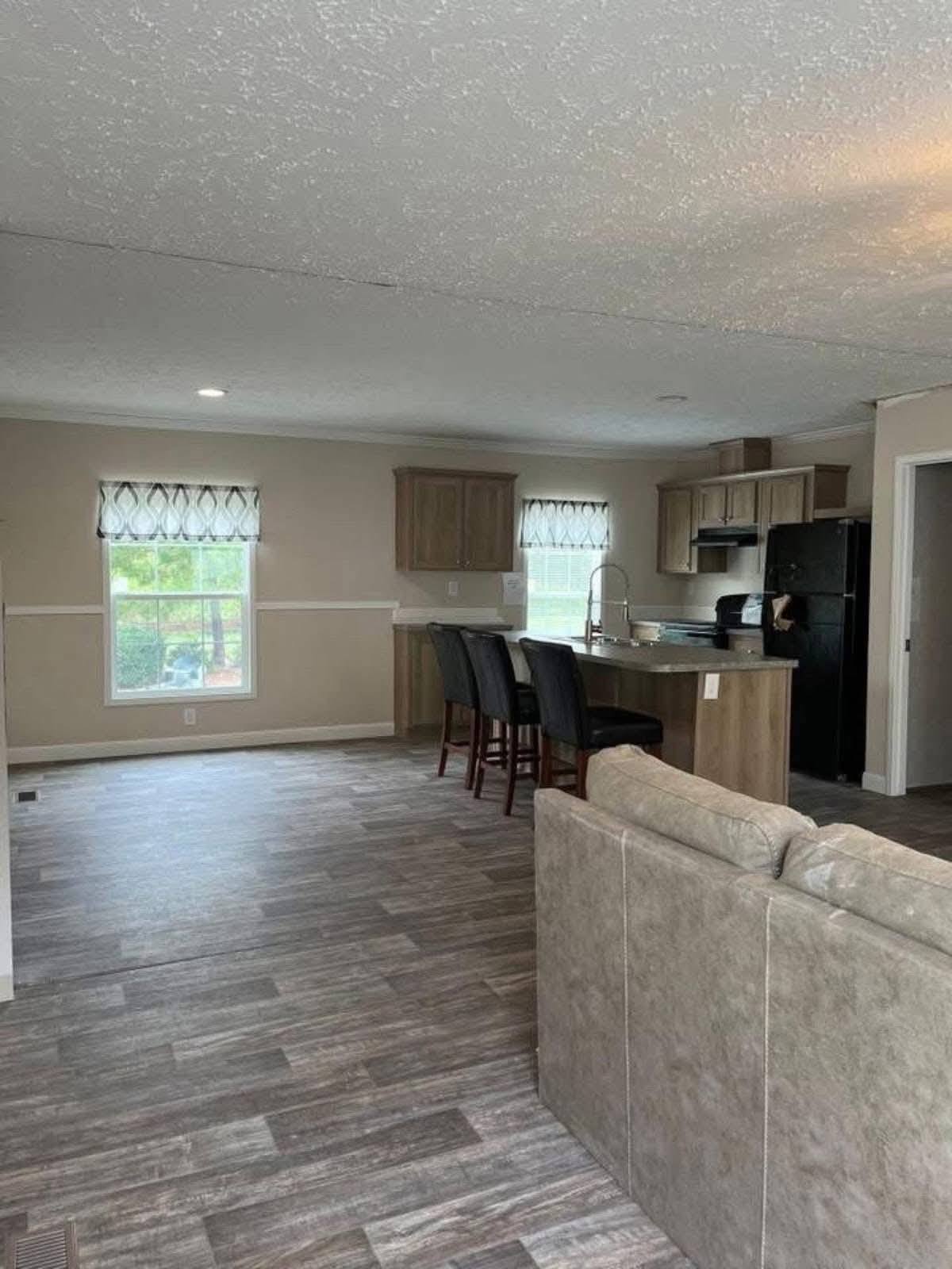 Open-plan living area with wood flooring, featuring a kitchen with light wood cabinets, black appliances, an island with bar stools, and a beige sofa.