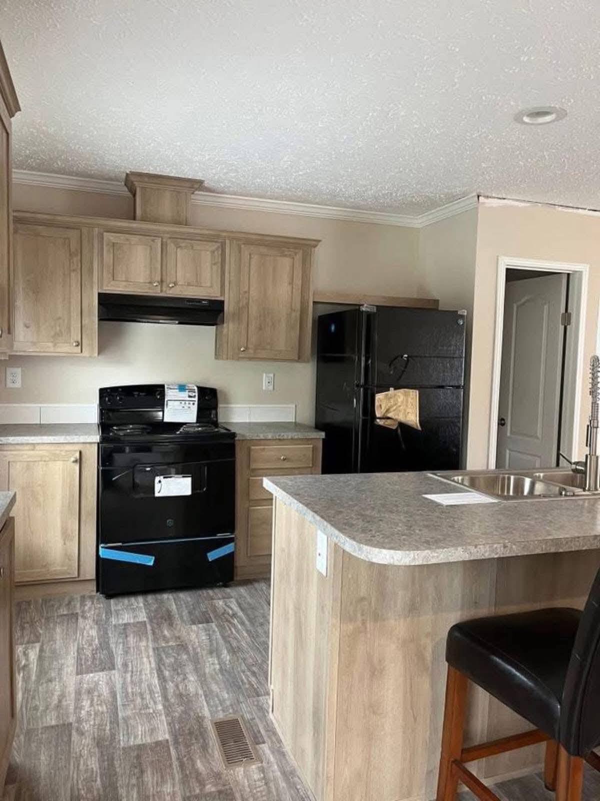 Compact, modern kitchen with light wood cabinets, black appliances, and gray countertops. Includes an island with a sink, creating a cozy and functional space.
