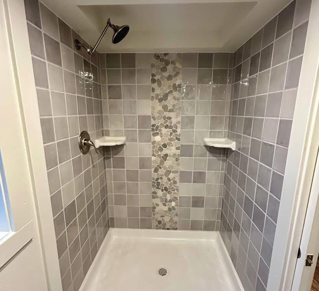 Modern shower with gray tiled walls and a pebble mosaic accent strip. Two corner shelves and a black showerhead are visible, creating a clean, serene look.